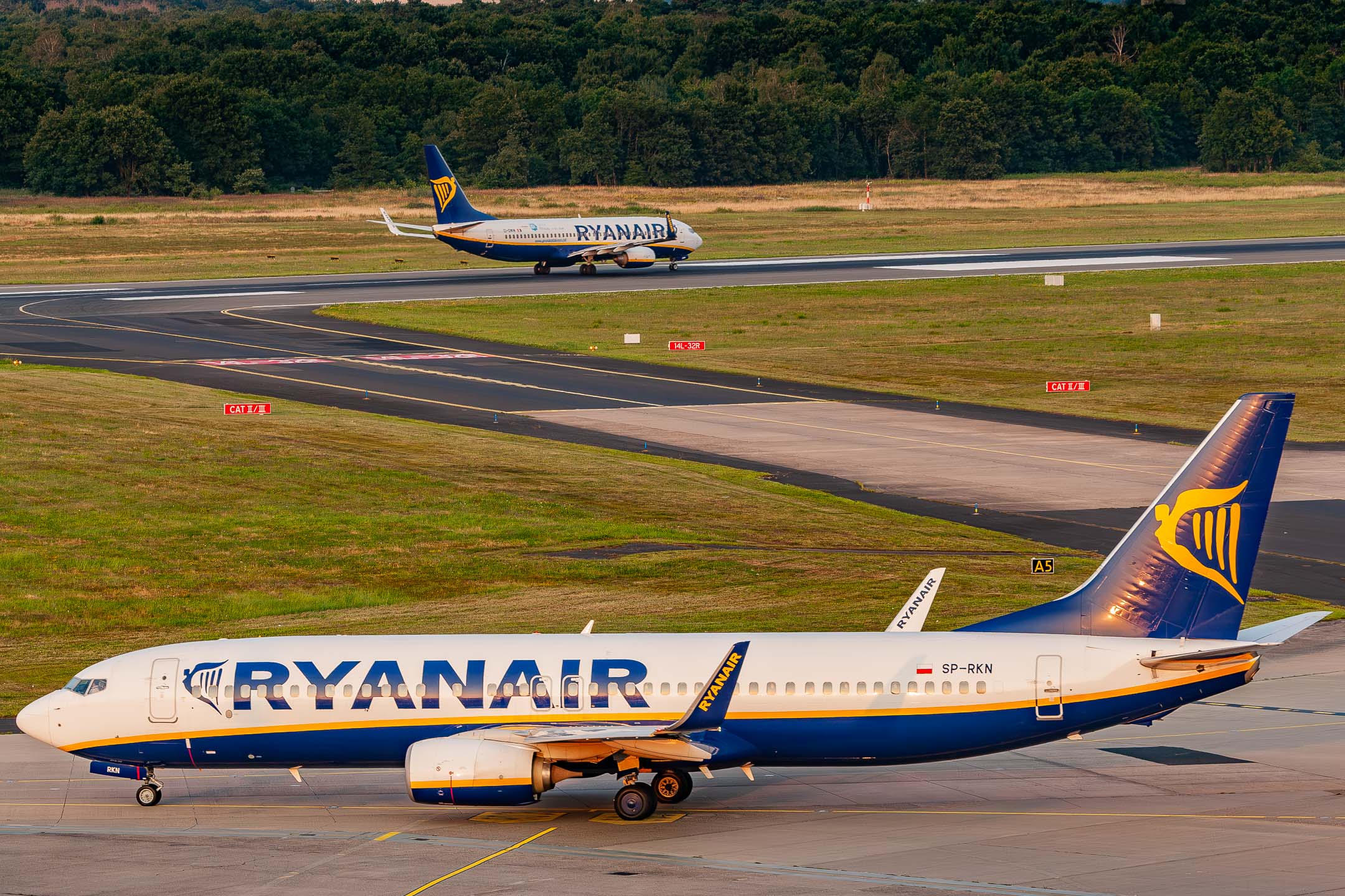 Ryanair Boeing 737-800 ("SP-RKN") taxiing in front of taxiway A5 while a company fellow accelerates on runway 14L at Köln Bonn Airport on a golden summer evening, July 2021.