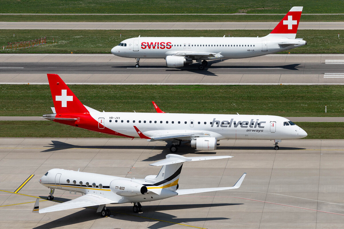 Helvetic Airways Embraer ERJ-190 ("HB-JVX") taxiing to the active runway 28 while Swiss Airbus A320 ("HB-IJL") accelerates for takeoff and a private Gulfstream V ("T7-BGD") is parked on the GAT apron on a very hot summer day at Zürich Airport, July 2023 